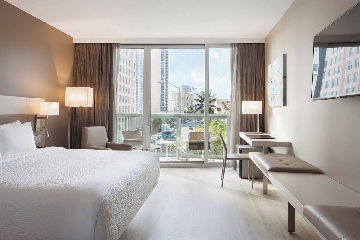 A modern hotel room with a large bed, a desk, a chair, and a balcony with a city view.