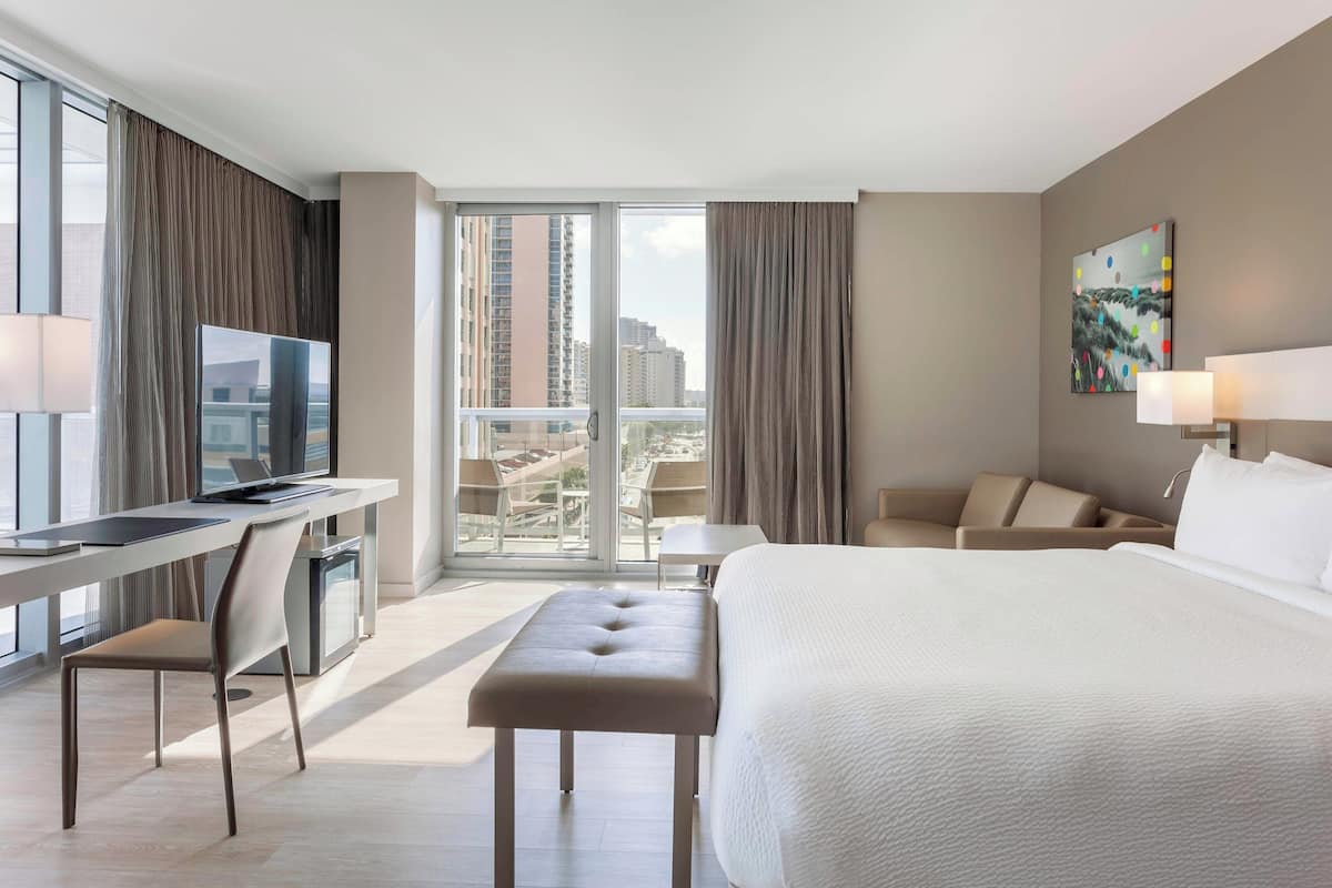 A modern hotel room with a large bed, a desk with a computer, a chair, and a view of the city through large windows.