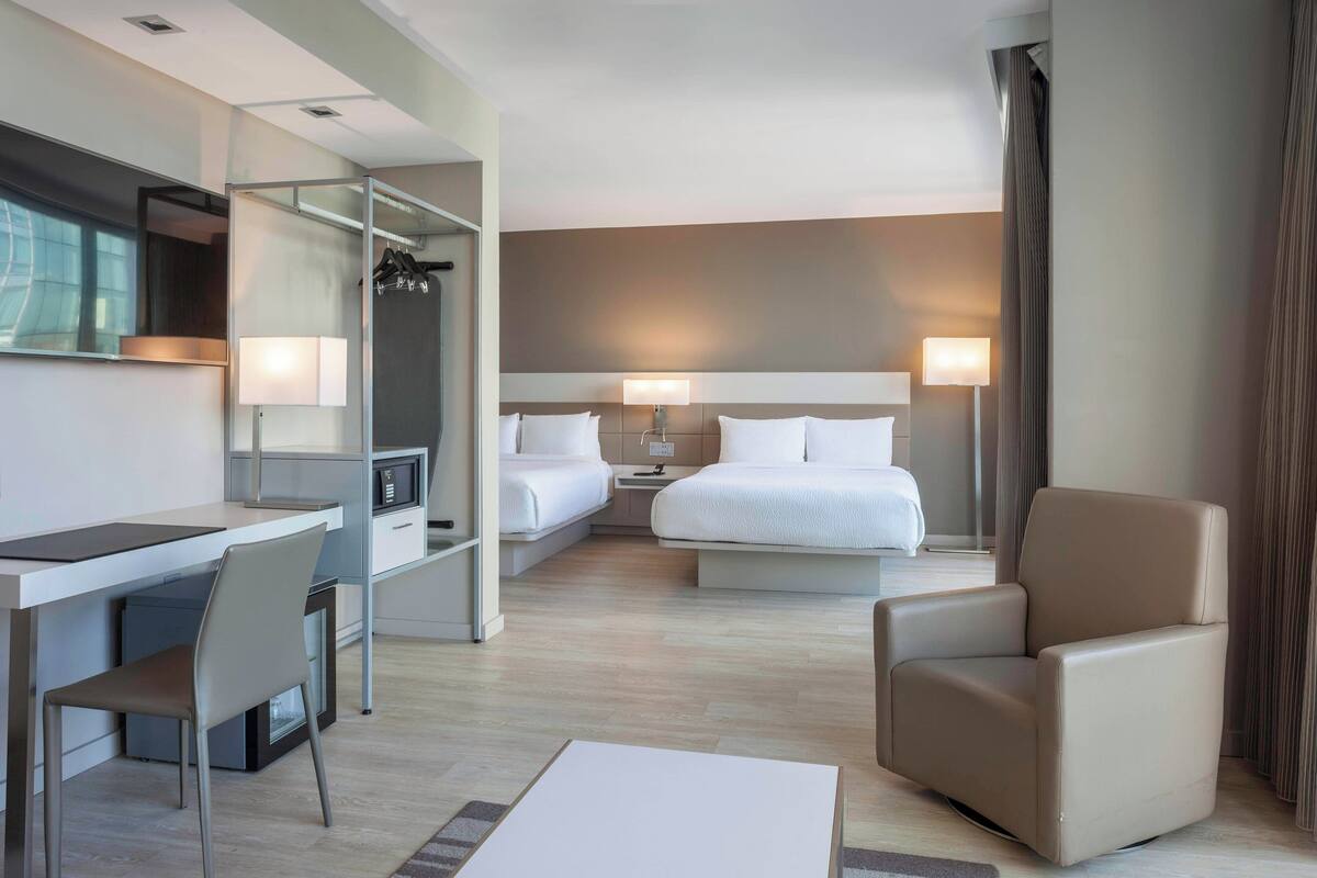 A modern hotel room with a desk, two beds, a chair, and a large mirror.