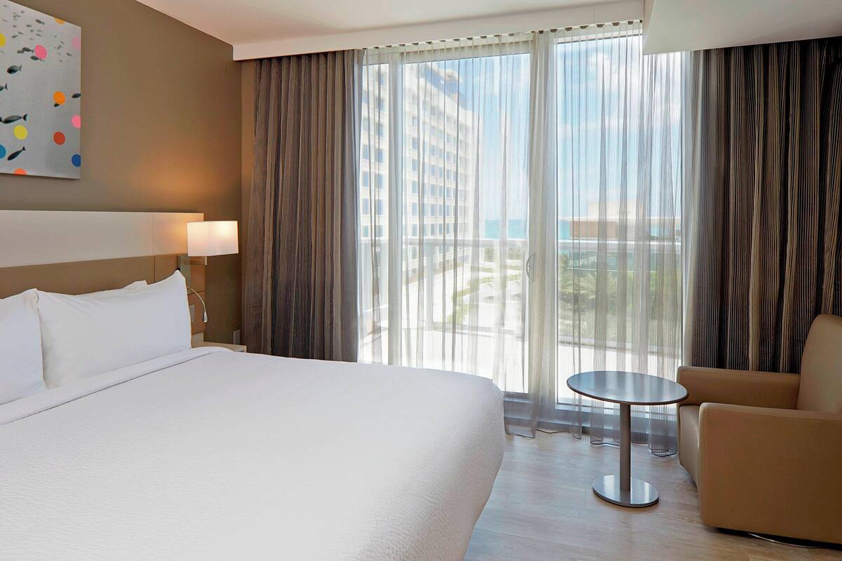 A hotel room with a large bed, a small round table, a sofa, and a view of the city through sheer curtains.