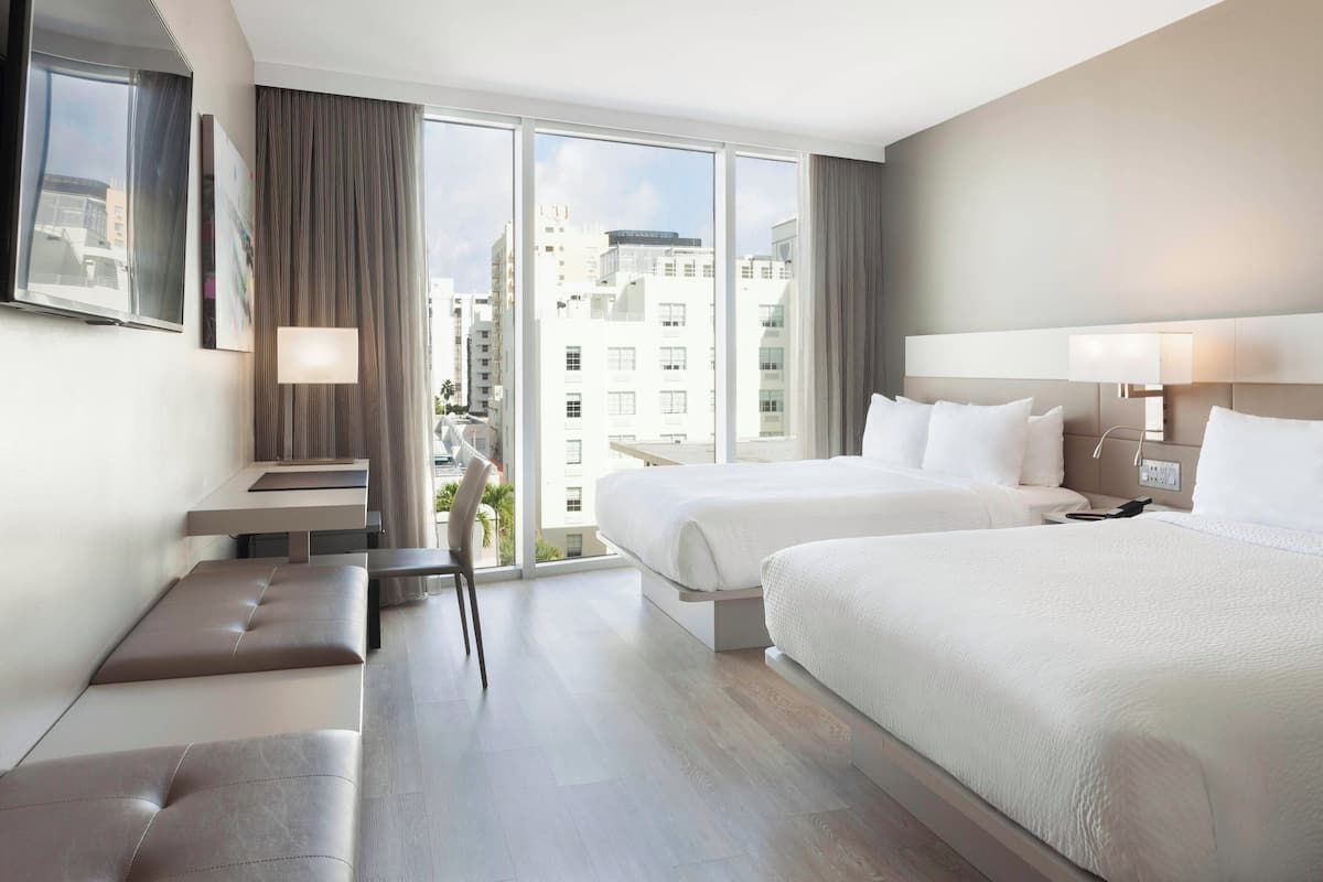 A modern hotel room with two beds, a seating area, and a large window offering a city view.