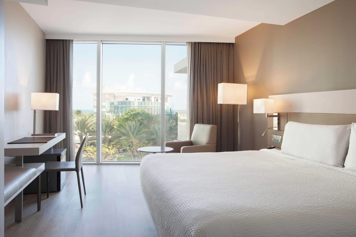 A modern hotel room with a large bed, a desk, a chair, and a view of the city through large windows.