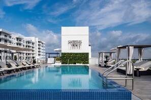 A rooftop pool area with lounge chairs, a pool with blue tiles, a white building with a mural, and a clear sky. - AC Hotel by Marriott Miami Beach (Miami Beach)