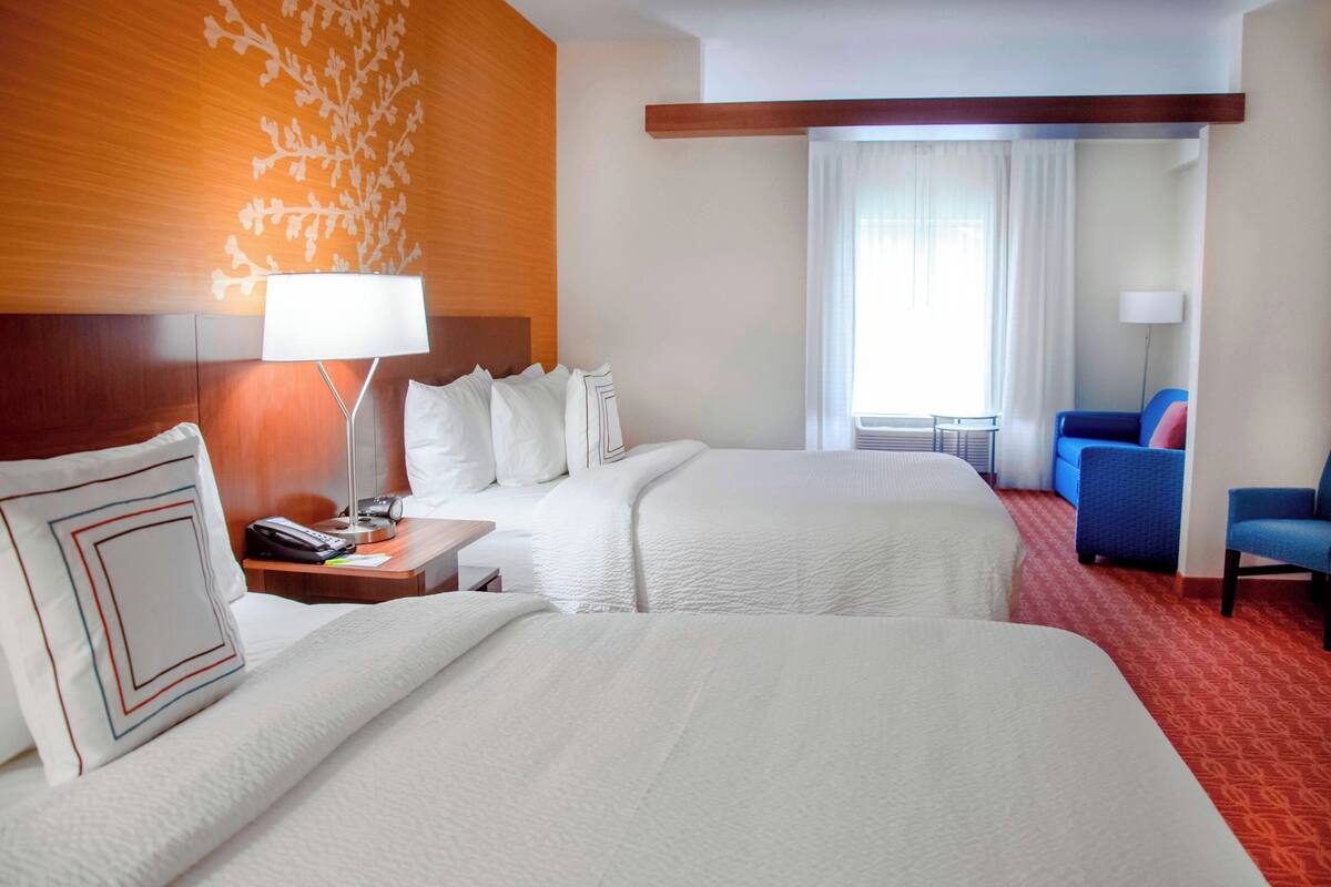 Habitación de hotel con dos camas, un escritorio con lámpara, una silla y un ventanal con cortinas.