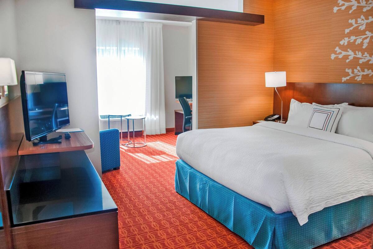 Una habitación de hotel con una cama grande, un escritorio con televisor de pantalla plana, una silla, una mesita y una l ámpara.