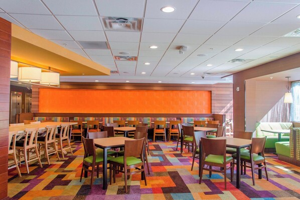 Un vestíbulo de hotel con una alfombra colorida, mesas y sillas de madera, y una pared grande de color naranja.