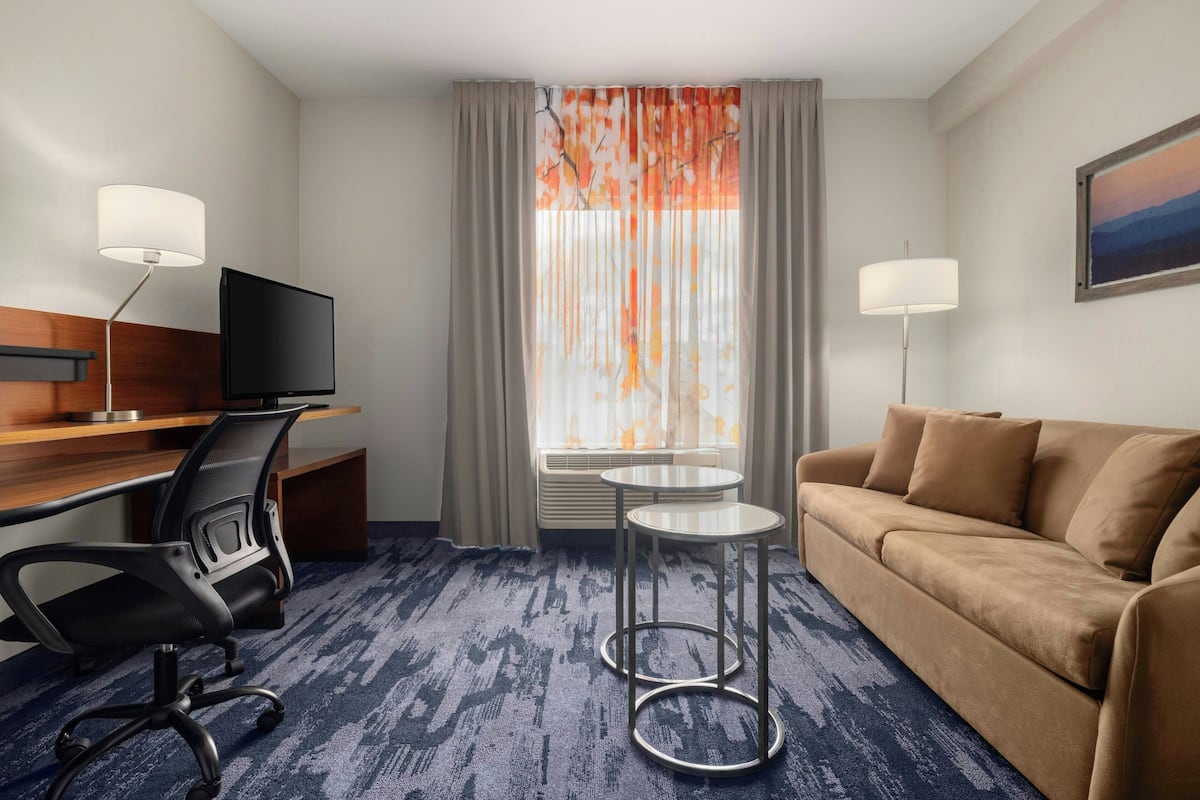 A hotel room with a desk, a television, a sofa, two side tables, and a window with curtains.