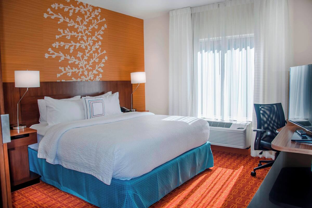 Una habitación de hotel con una cama grande, dos lámparas de noche, un escritorio con una computadora y una ventana con cortinas translúcidas.