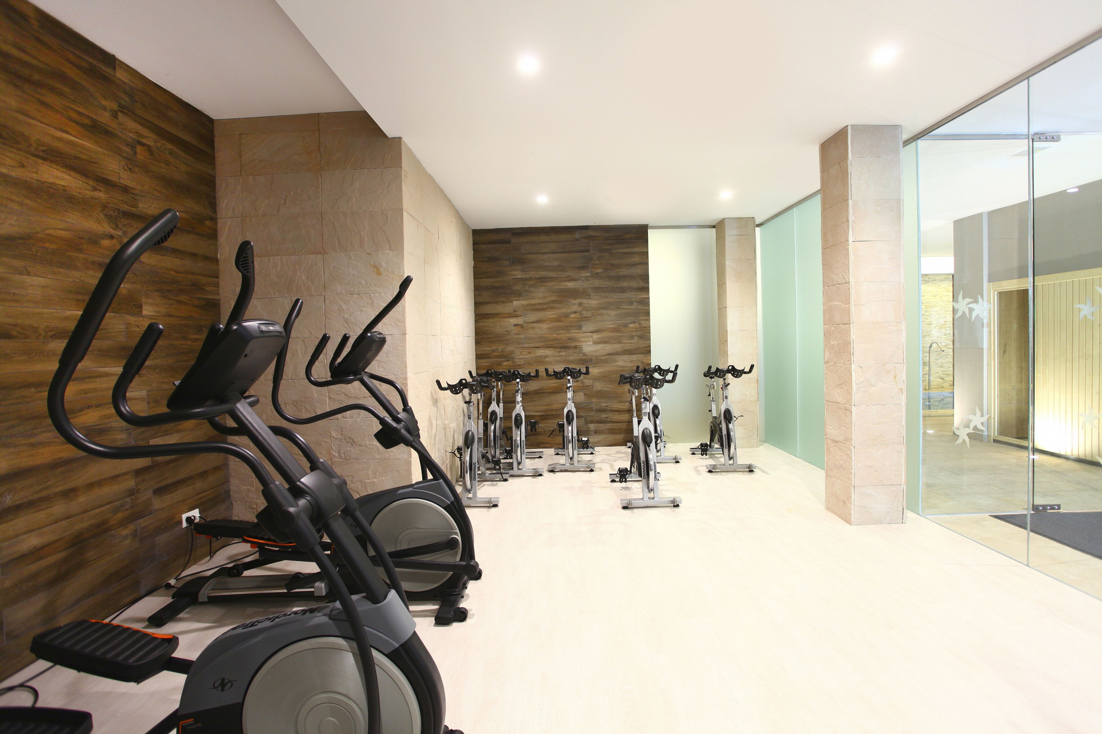 A gym with multiple stationary bikes arranged in rows, a wooden wall, and a glass door leading to another room.