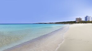 A beach with clear turquoise water, sandy shore, and buildings in the background. - Iberostar Waves Cala Millor -Adults Only (Sant Llorenc des Cardassar)