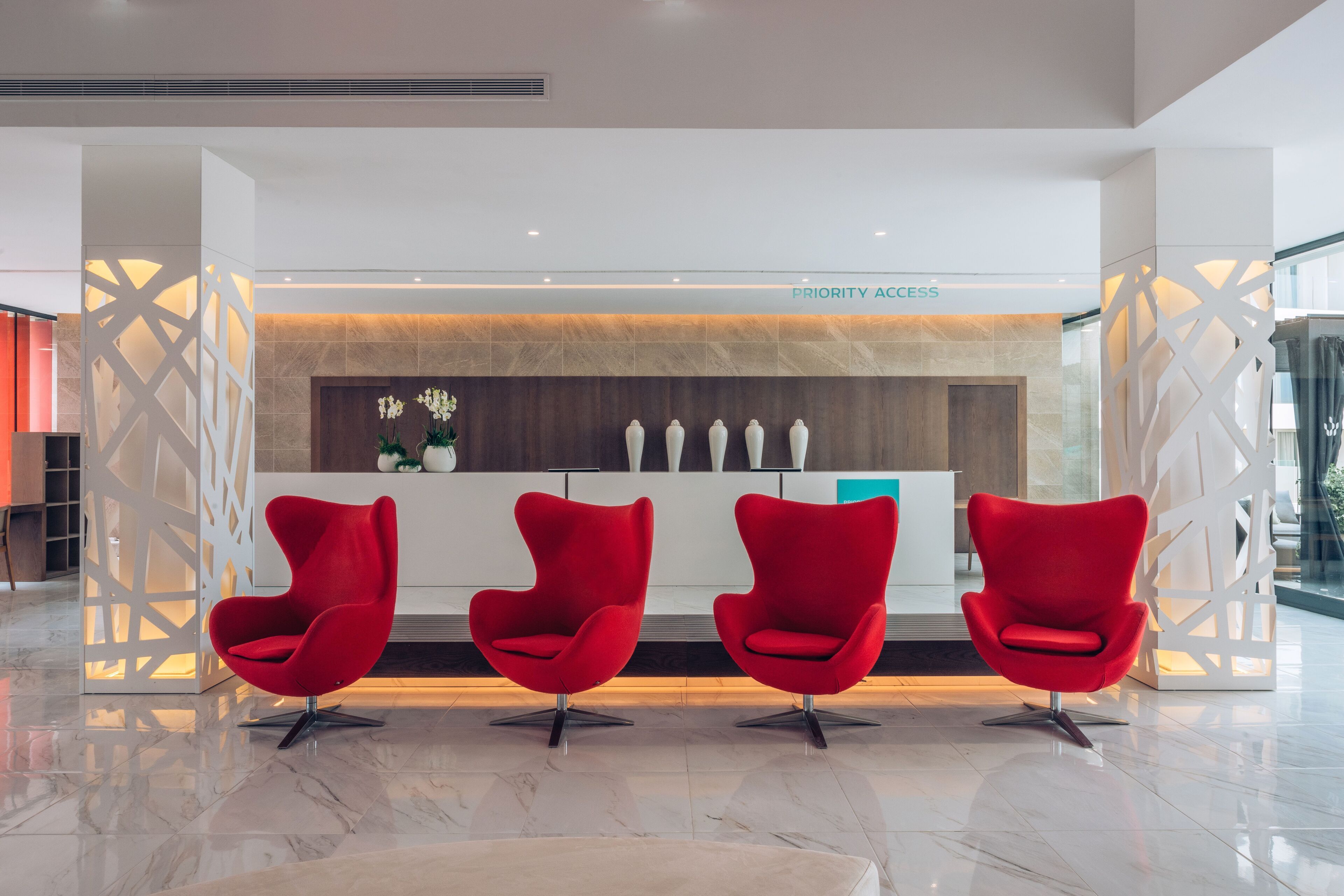 Un espace d’accueil au design moderne, comprenant un comptoir d’accueil, trois chaises rouges et un panneau indiquant « Accès prioritaire ».