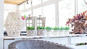 Una habitación con un ventanal grande, una silla de mimbre y varias macetas con plantas en un estante.