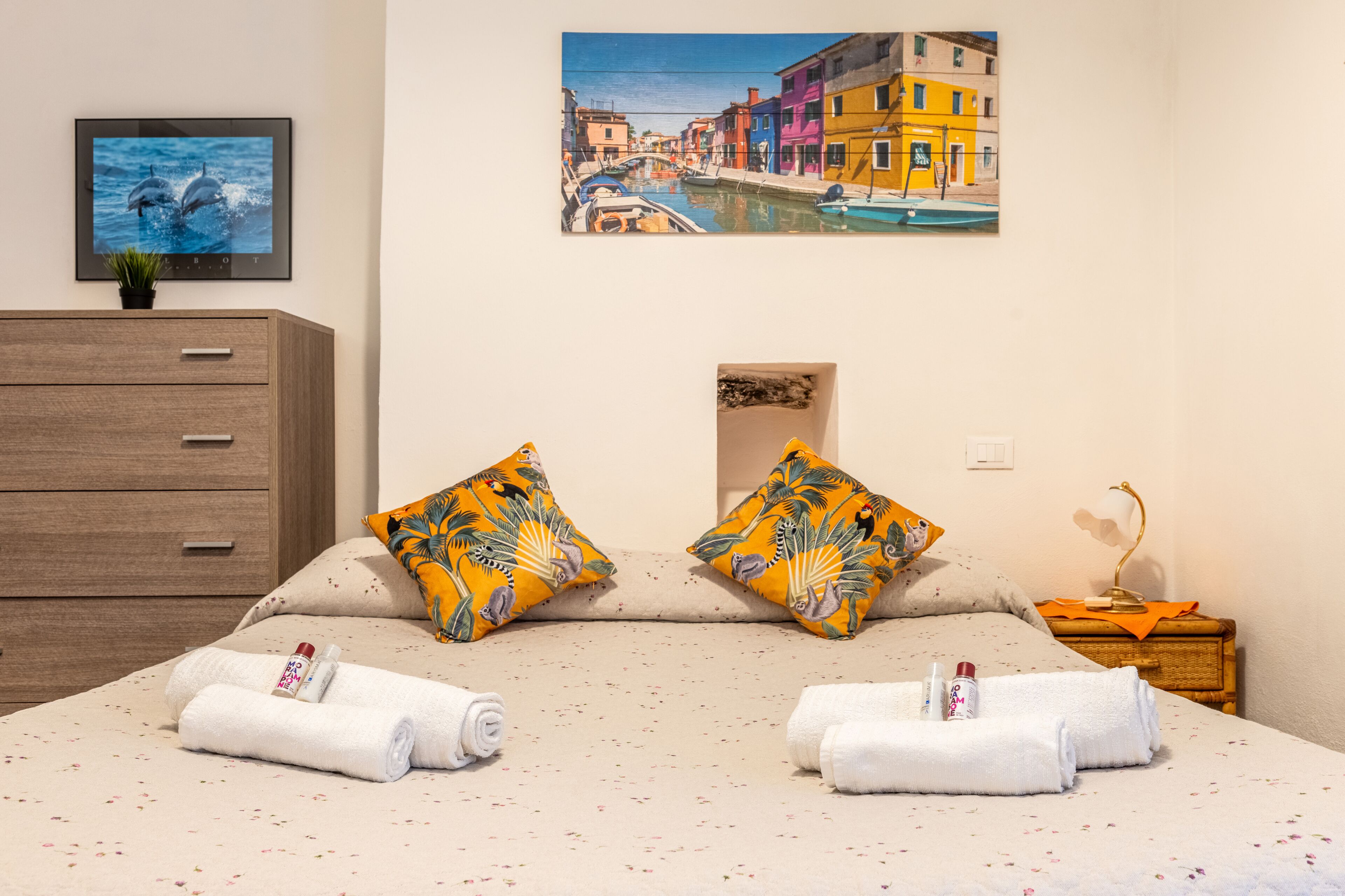 Une chambre d’hôtel comprenant un lit, deux oreillers, une table de chevet, une lampe, un tableau, une télévision et une commode en bois.