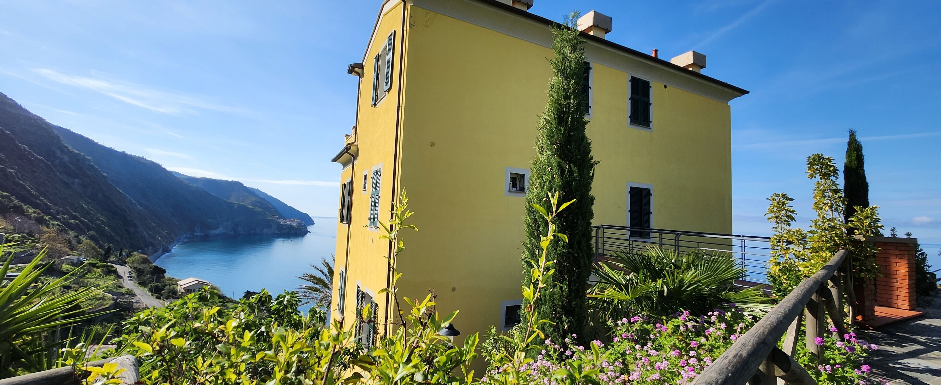 A yellow building with a balcony overlooking a coastal area with mountains and the sea.