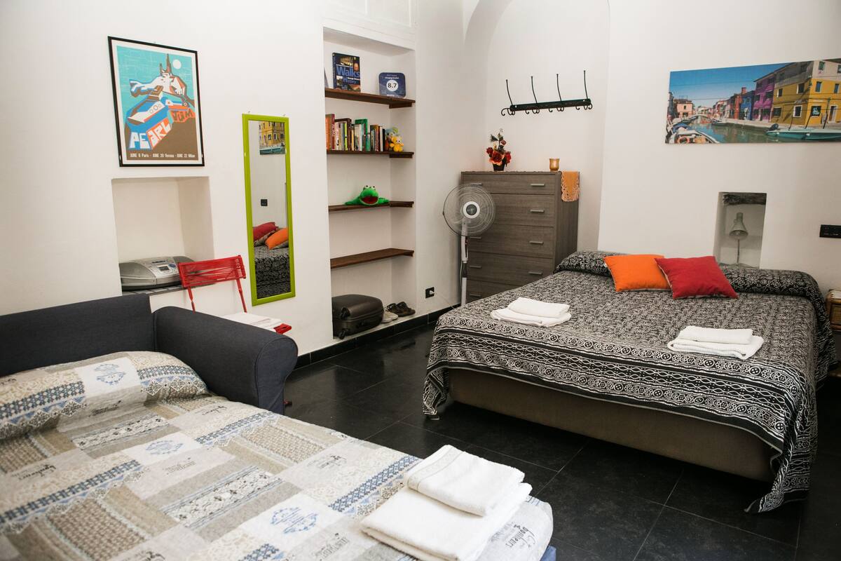 A bedroom with a bed, a sofa, a dresser, a mirror, a fan, and a bookshelf.