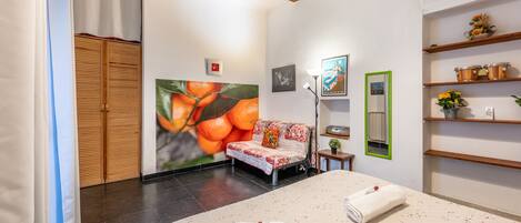 A hotel room with a bed, a sofa, a wooden wardrobe, a wall with a fruit painting, and shelves with decorative items.
