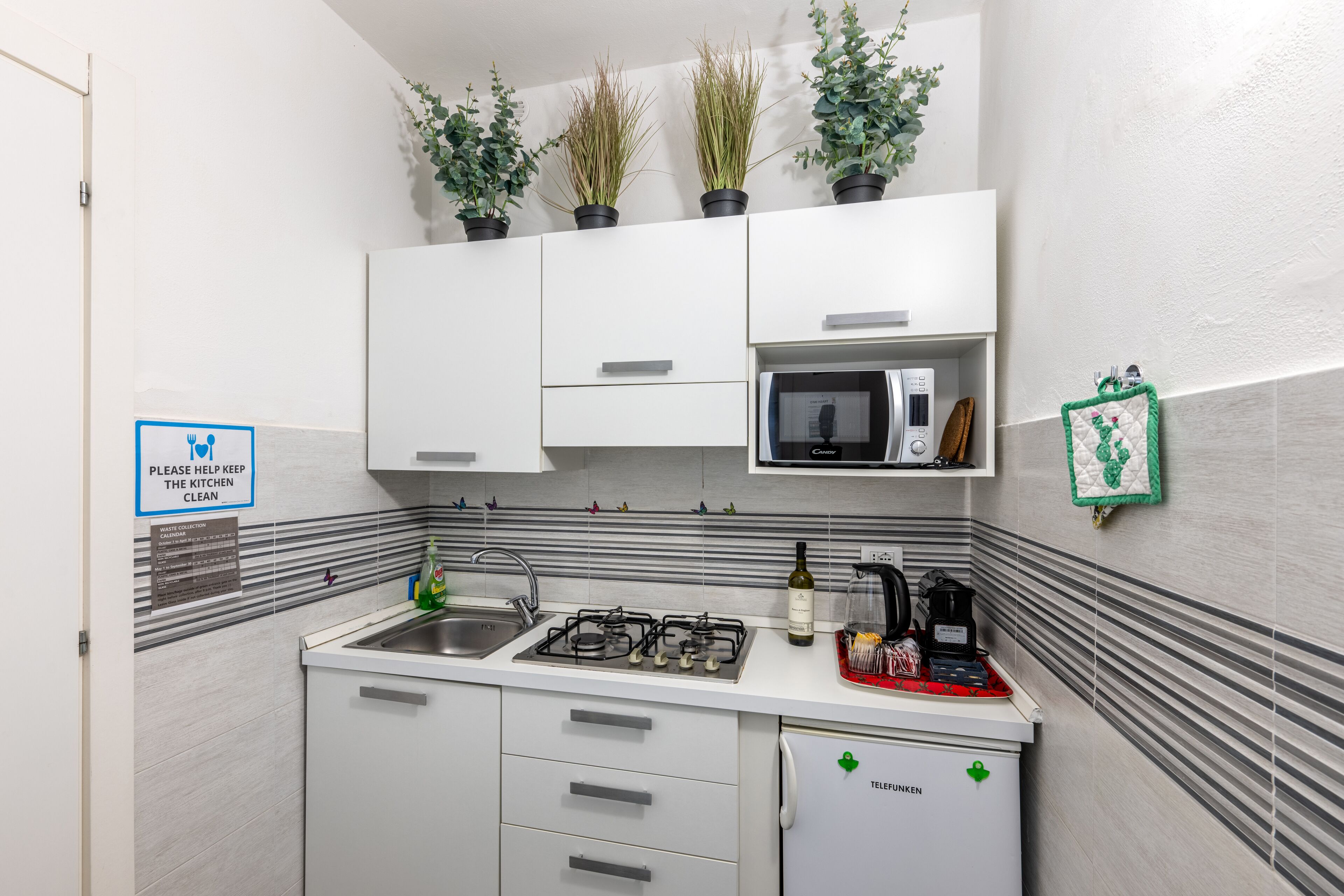 A compact kitchen with white cabinets, a microwave, a gas stove, and a refrigerator. There are potted plants on the wall, a sink, and a sign requesting kitchen cleanliness.