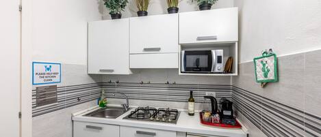 A compact kitchen with white cabinets, a microwave, a gas stove, and a refrigerator. There are potted plants on the wall, a sink, and a sign requesting kitchen cleanliness.
