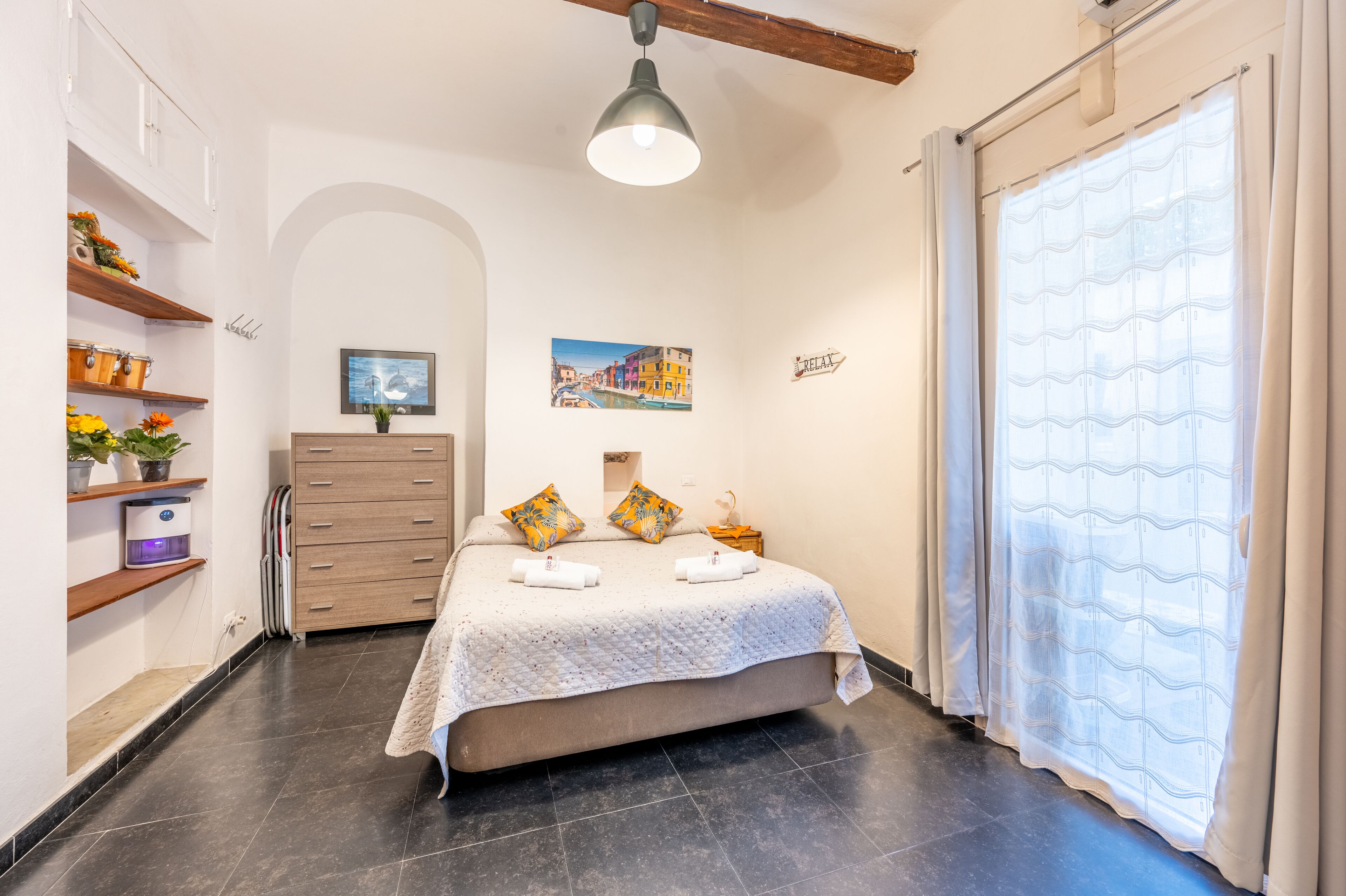 A bedroom with a bed, a dresser, shelves with plants, a painting, and a window with curtains.