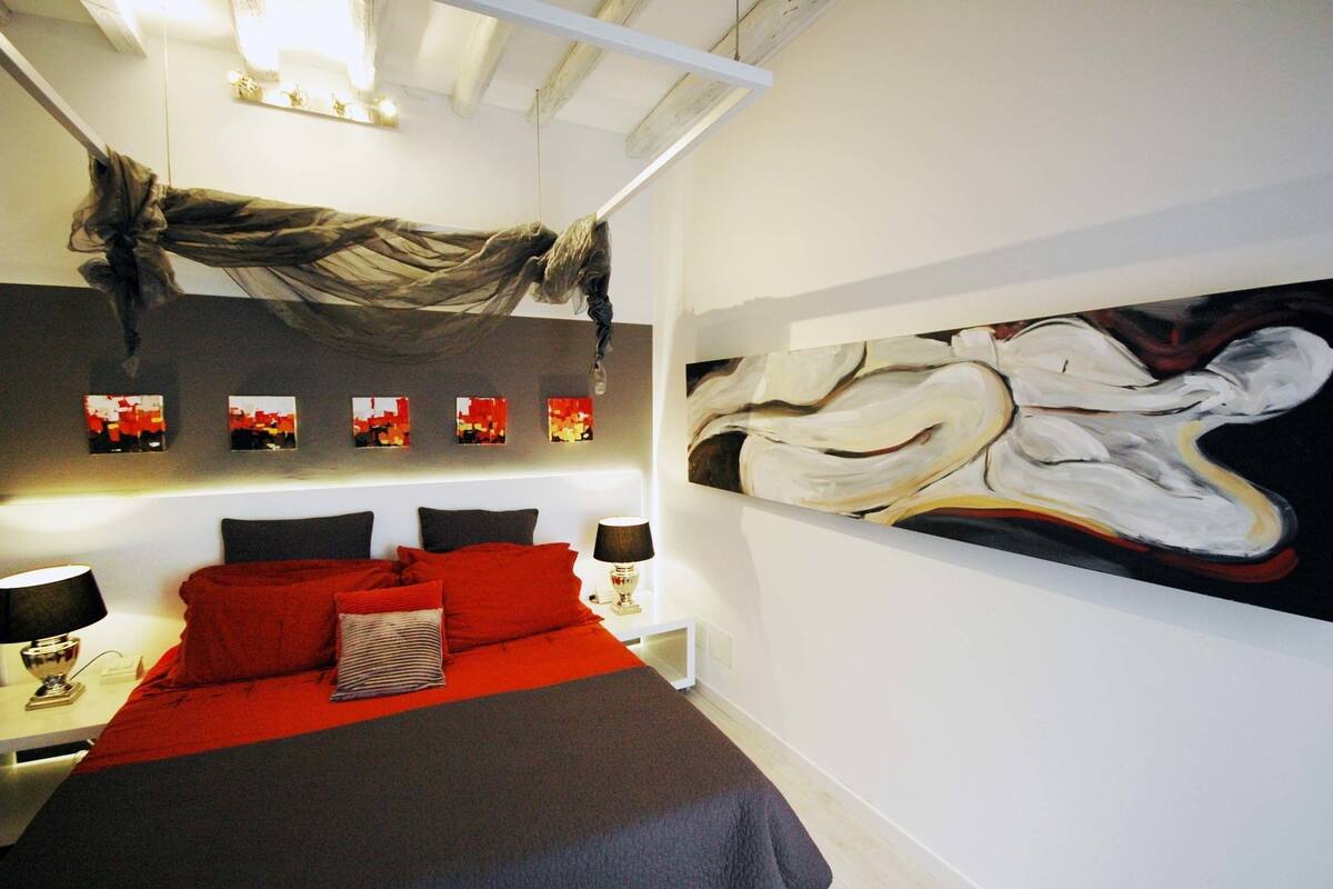 A modern hotel room with a large bed, a headboard with multiple lights, a wall-mounted artwork, and a bedside table with a lamp.