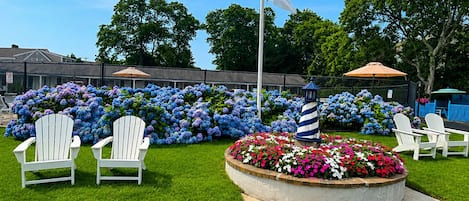 A flagpole with an American flag, a circular flower bed with pink and white flowers, and white Adirondack chairs.