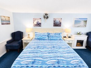 A hotel room with a bed, two nightstands, a chair, a fireplace, and wall art. - A Beach Breeze Inn (West Harwich)