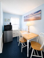 Una habitación compacta con kitchenette, una mesa de comedor con cuatro sillas, una lámpara y un cuadro en la pared.