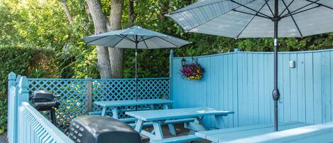 Un espace extérieur aménagé avec des sièges bleus, des tables de pique-nique et des parasols, entouré d’une clôture en bois et d’arbres.