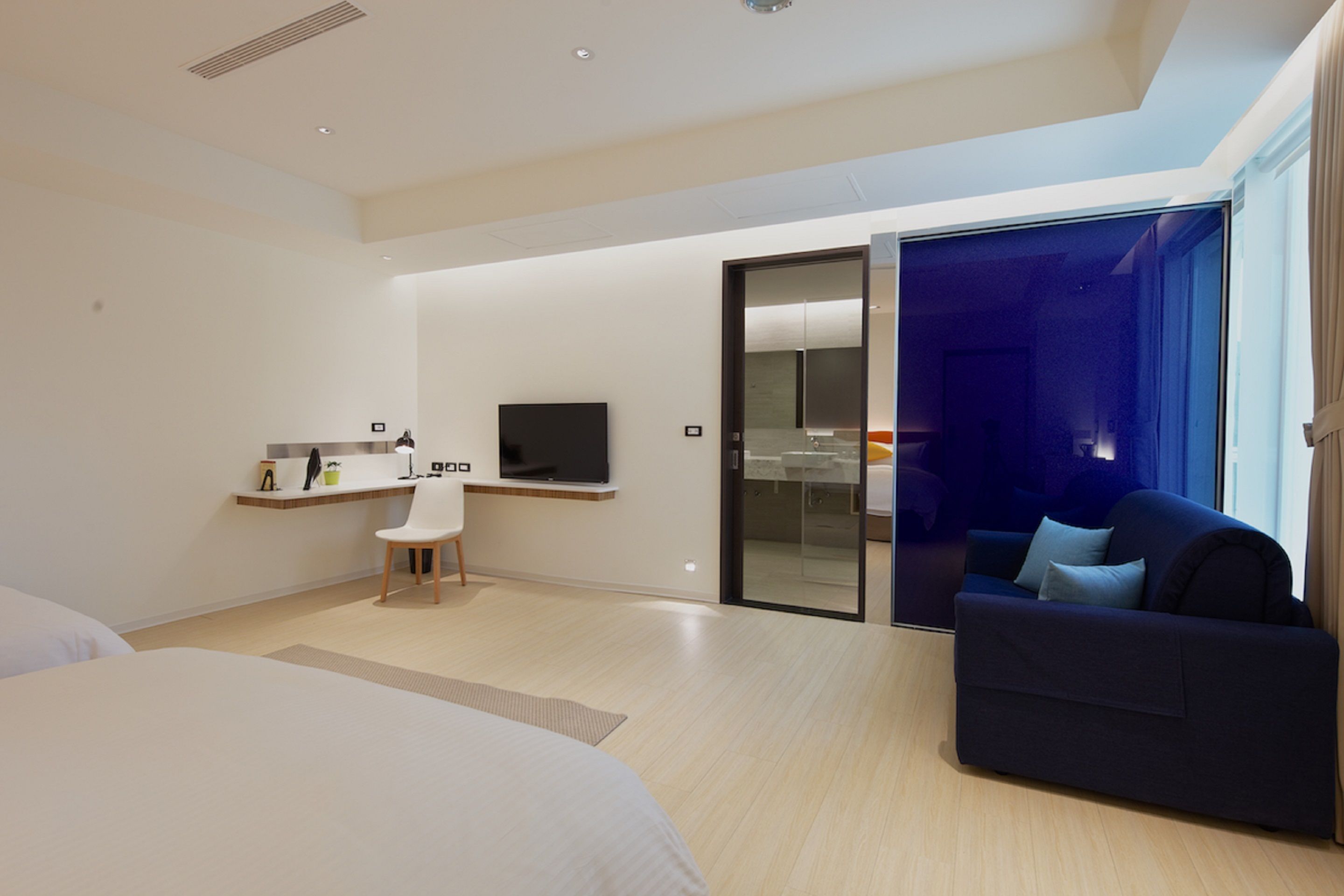 A modern hotel room with a bed, a desk with a chair, a flat-screen TV, and a dark-colored sofa.