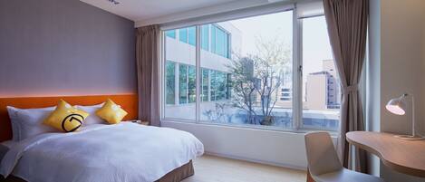 A hotel room with a large bed, a desk with a lamp, and a view of buildings through the window.