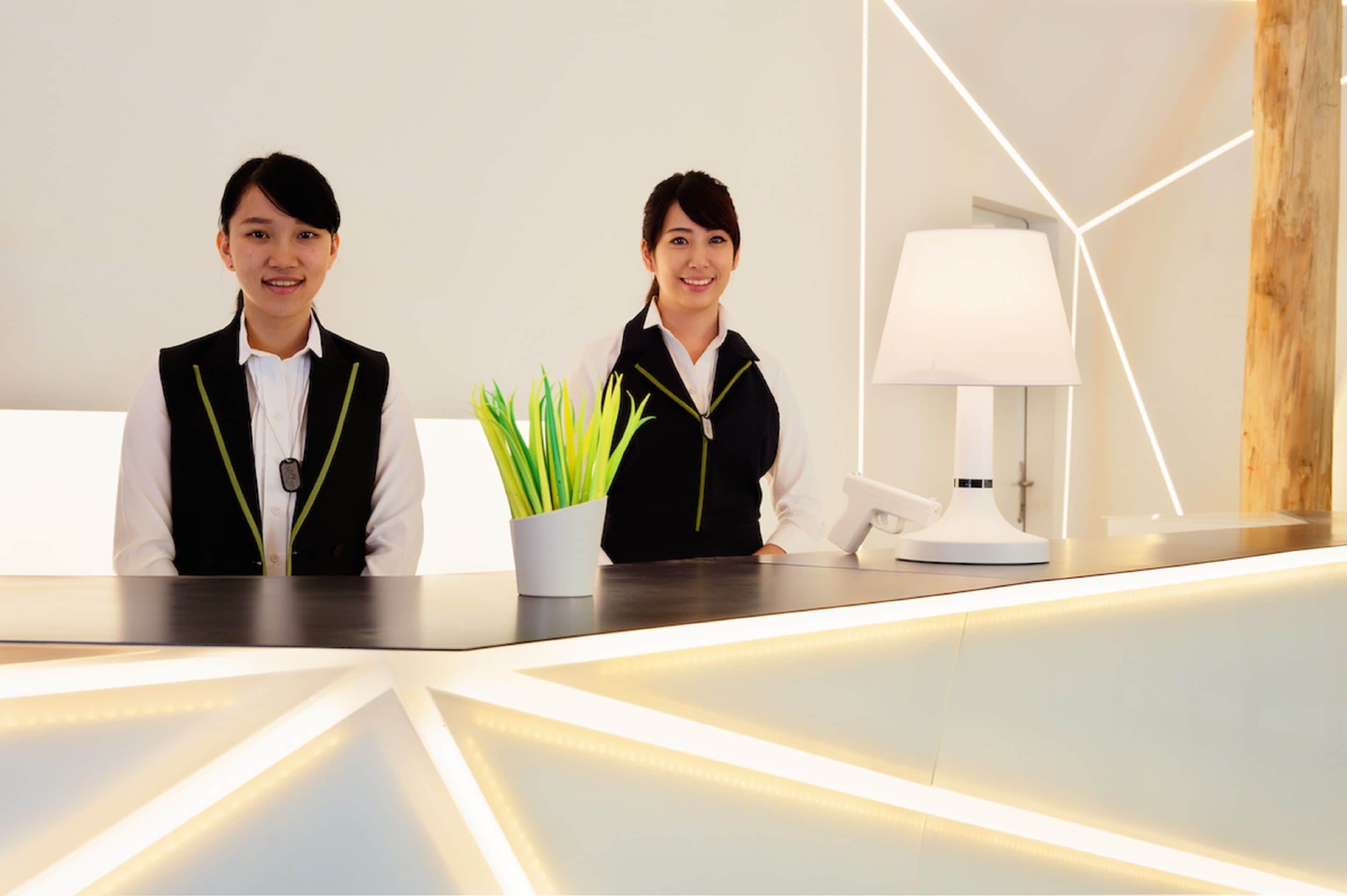 Two receptionists at a modern reception desk with a potted plant and a lamp.