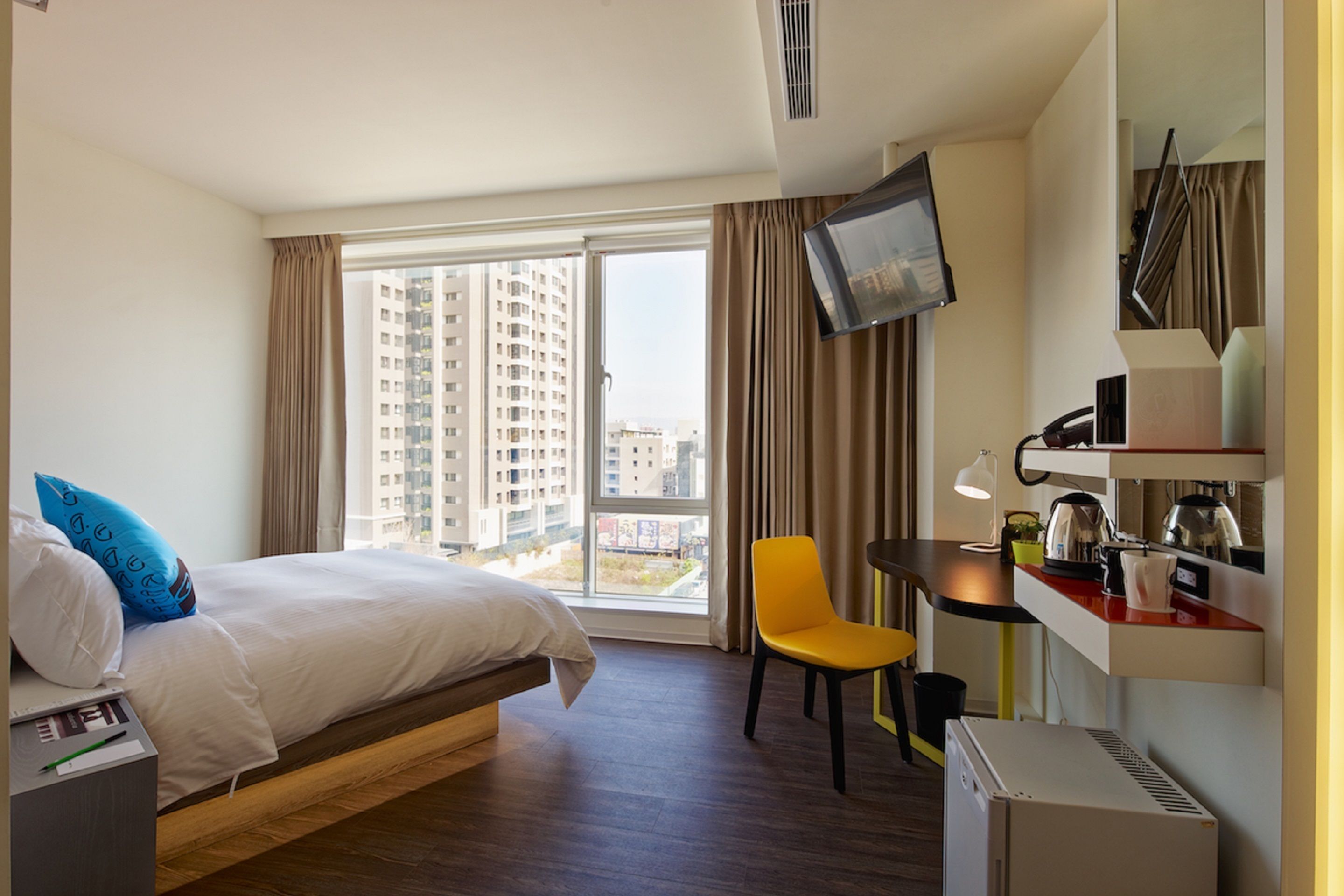 A hotel room with a bed, a desk, a chair, a TV, and a view of tall buildings.