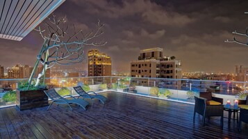 A rooftop terrace with lounge chairs, a decorative tree, and a view of city buildings at night.