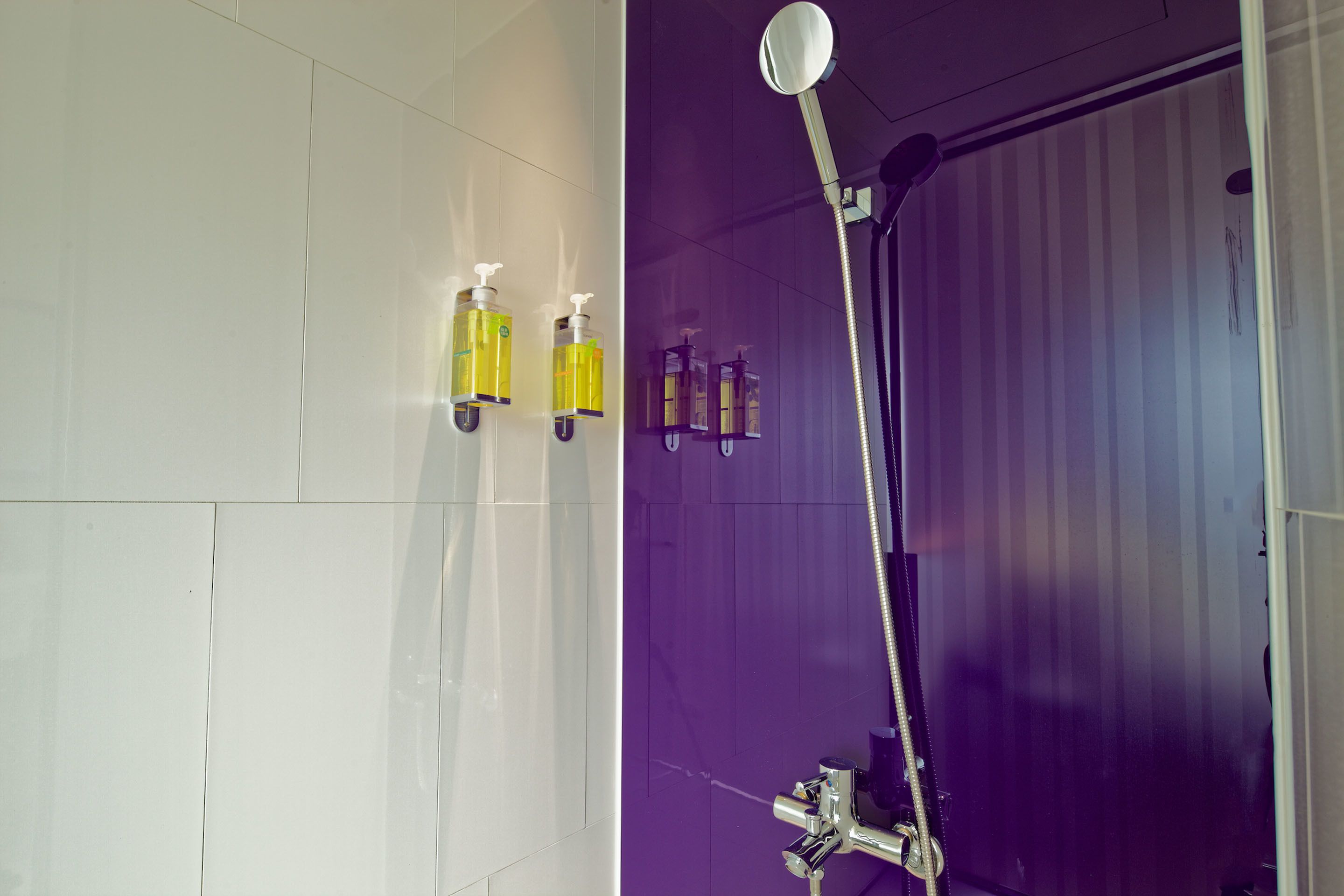 Um banheiro com paredes revestidas de azulejos brancos, box de chuveiro roxo e duas garrafas de shampoo amarelas na parede.