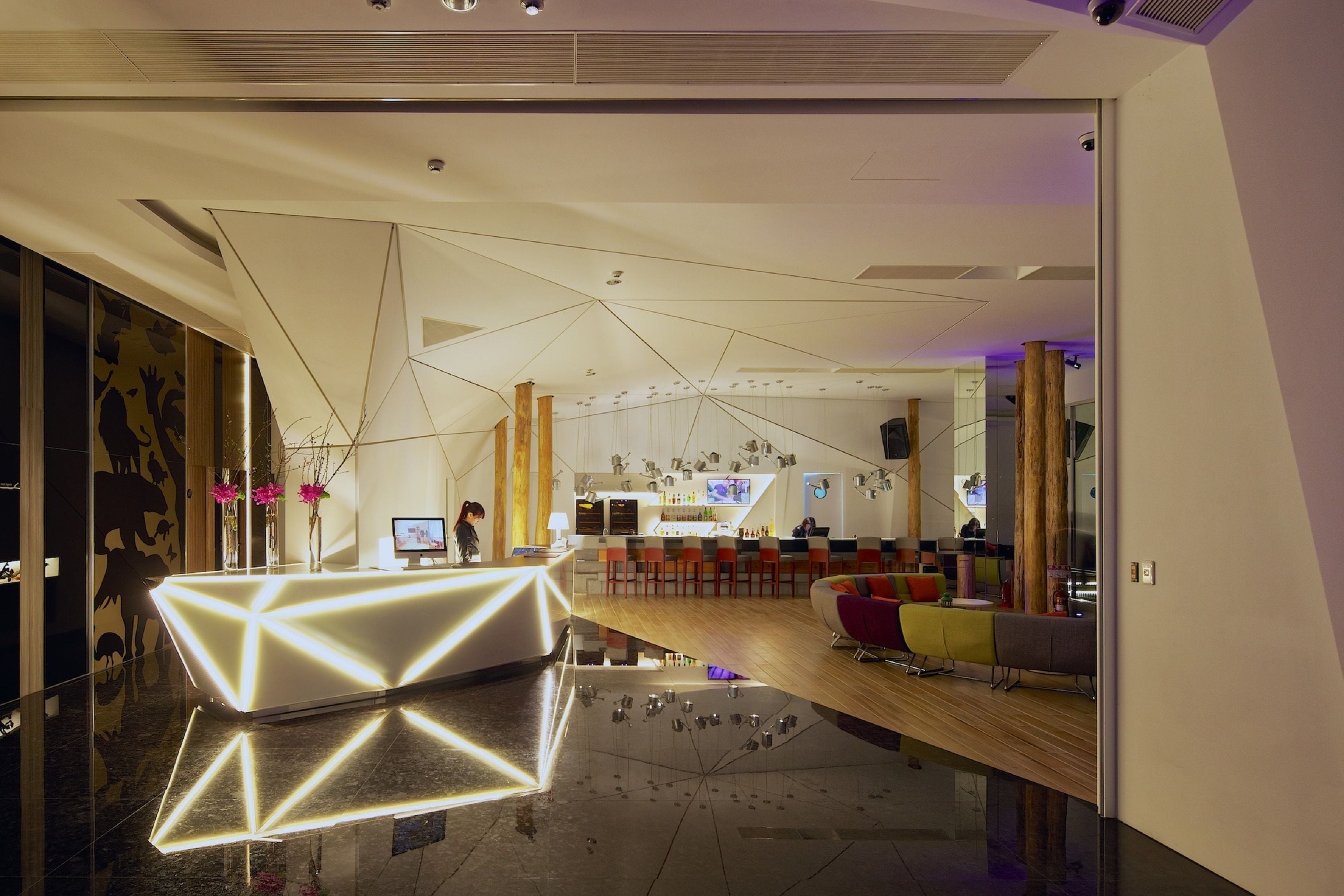 A modern hotel lobby with a geometric reception desk, a bar area, and a seating space with colorful chairs.