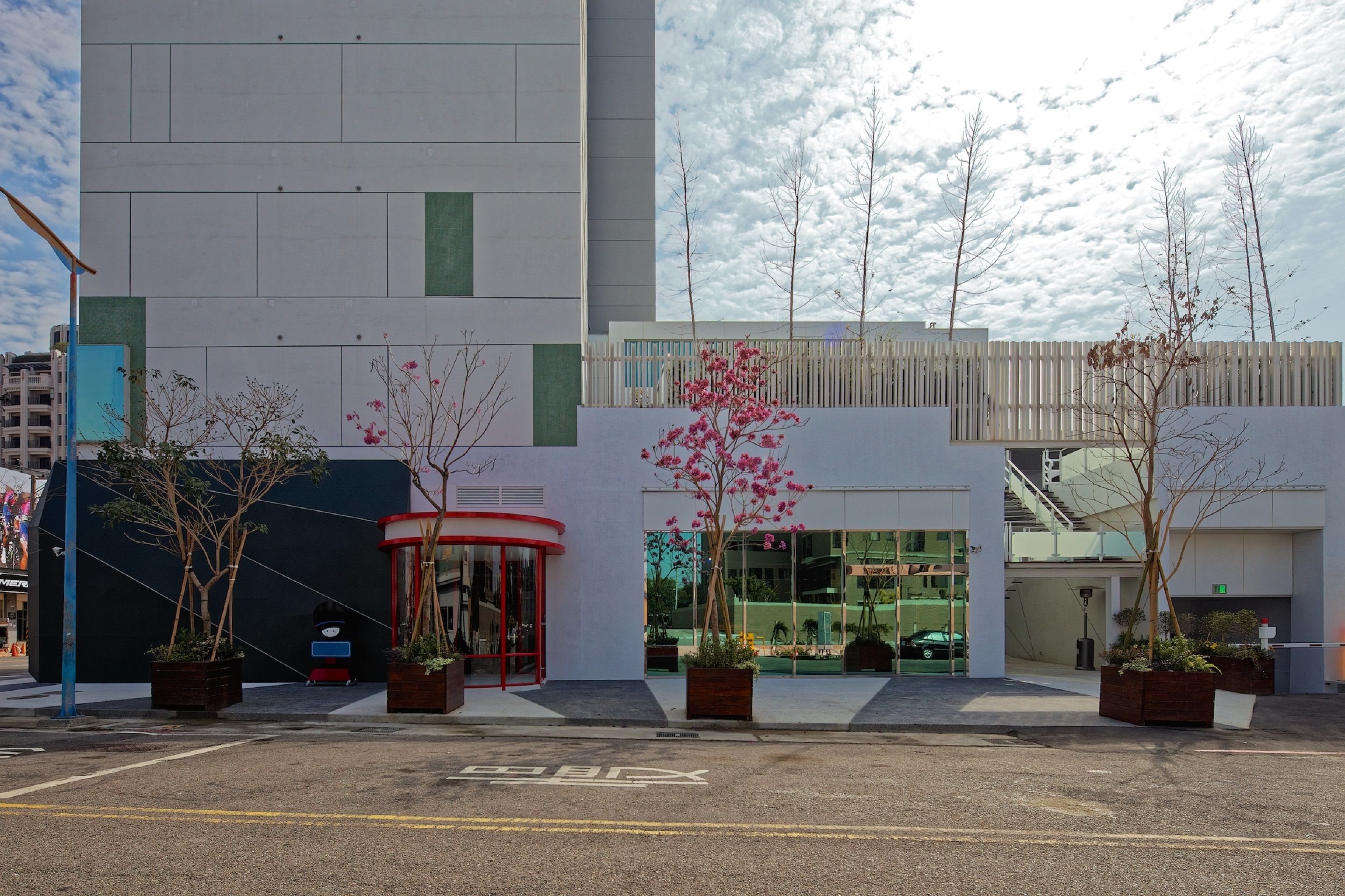 A modern building with a curved entrance, large windows, and a parking area with potted plants.