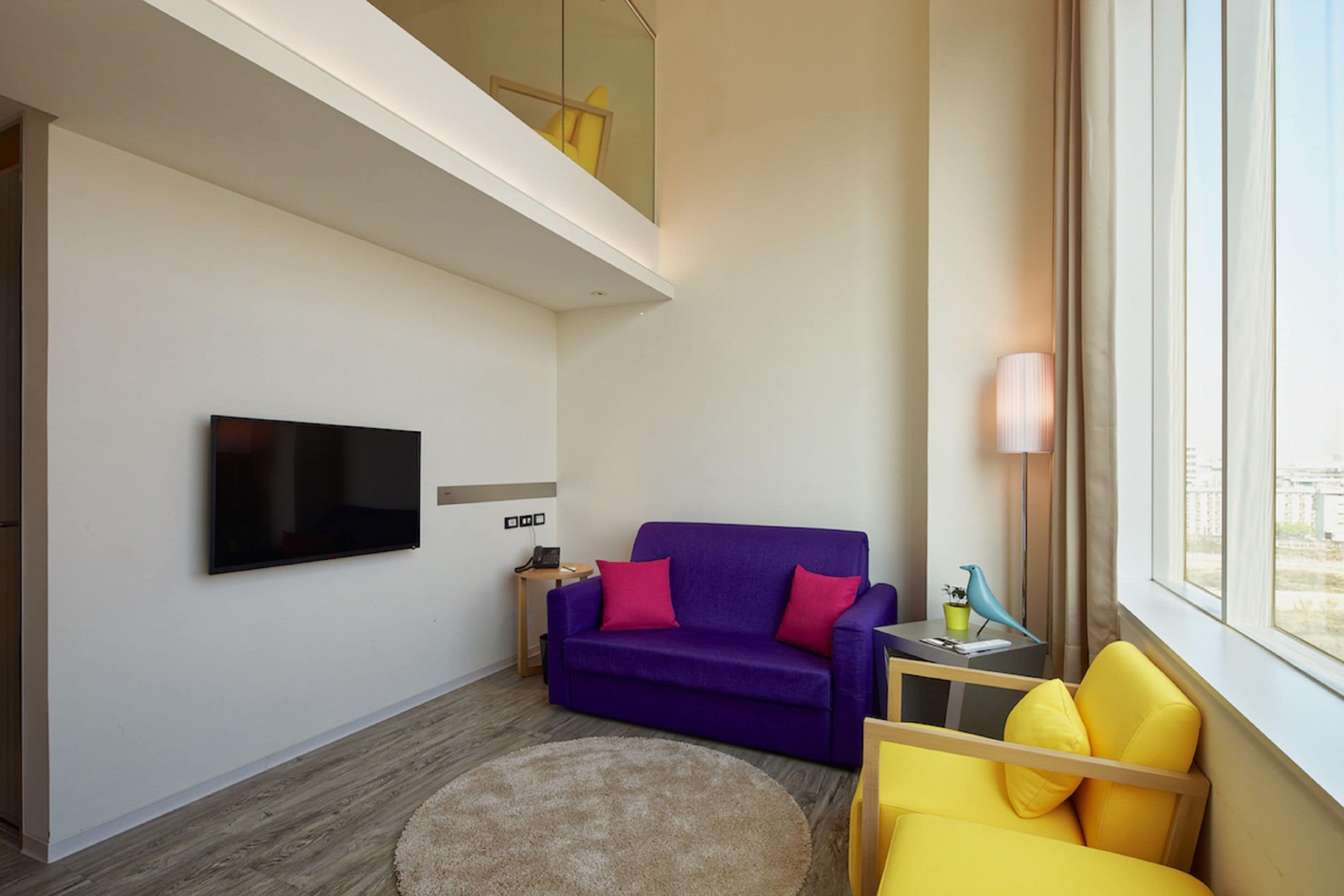 A modern living room with a flat-screen TV, a purple sofa, a yellow armchair, and a round rug.