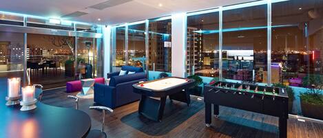 A modern lounge area with a foosball table, comfortable seating, and large windows offering city views.