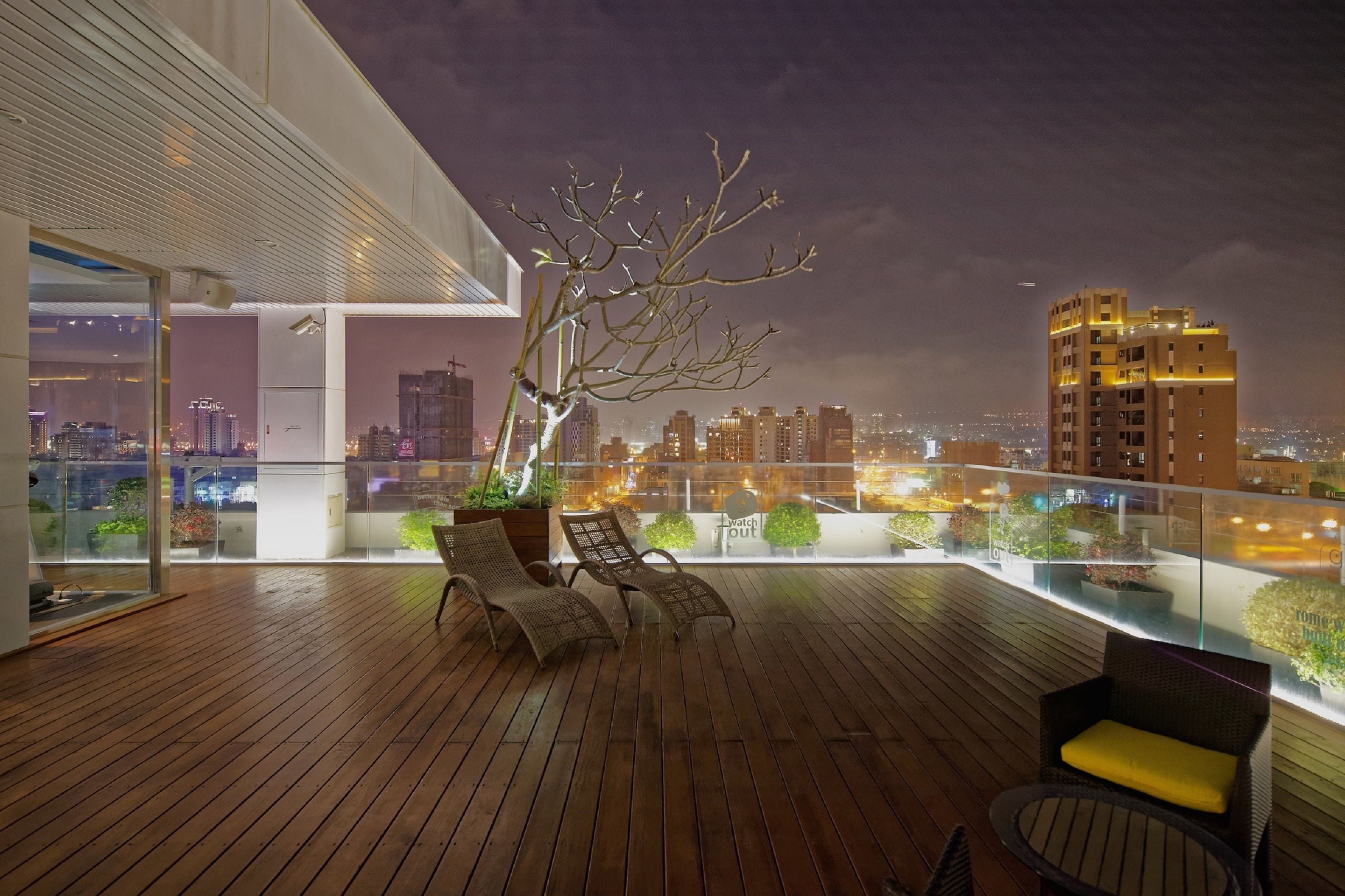 Um deck de madeira com duas cadeiras de vime e uma mesa pequena, com vista para a cidade à noite.