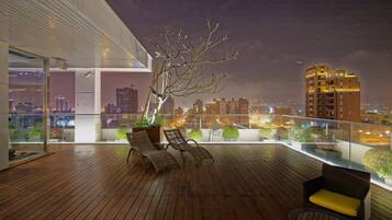 A wooden deck with two wicker chairs and a small table, overlooking a cityscape at night.