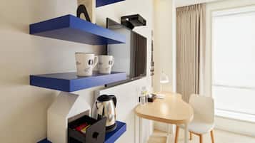 A modern hotel room with blue shelving units, a coffee maker, a kettle, a table with a chair, and a large window with curtains.