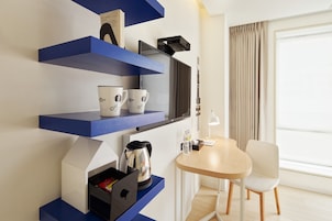 A modern hotel room with blue shelving units, a coffee maker, a kettle, a table with a chair, and a large window with curtains.