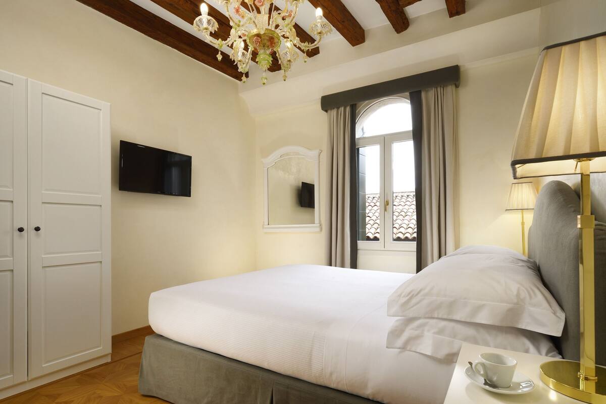 A bedroom with a bed, a television mounted on the wall, a chandelier, a window with curtains, and a bedside table with a cup and saucer.