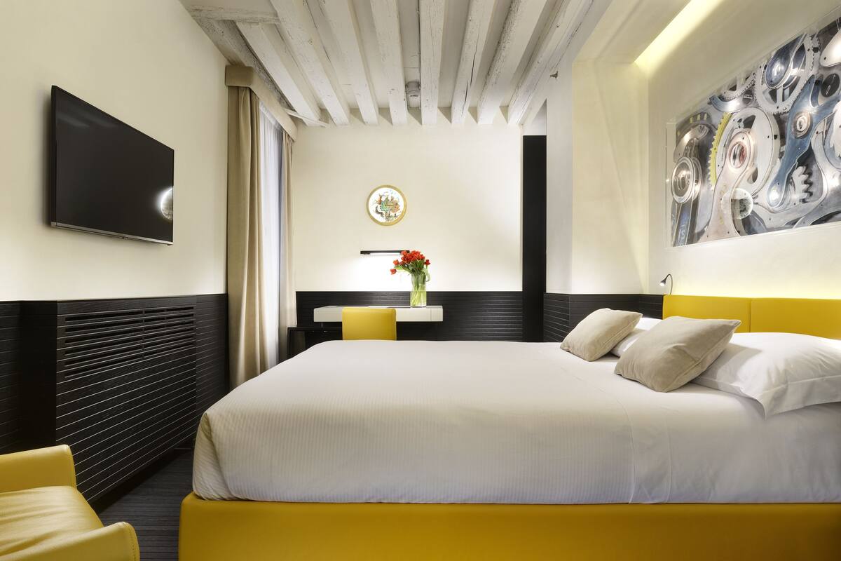A modern hotel room with a large bed, a flat-screen TV, a small table with a vase of flowers, and a unique wall art piece.