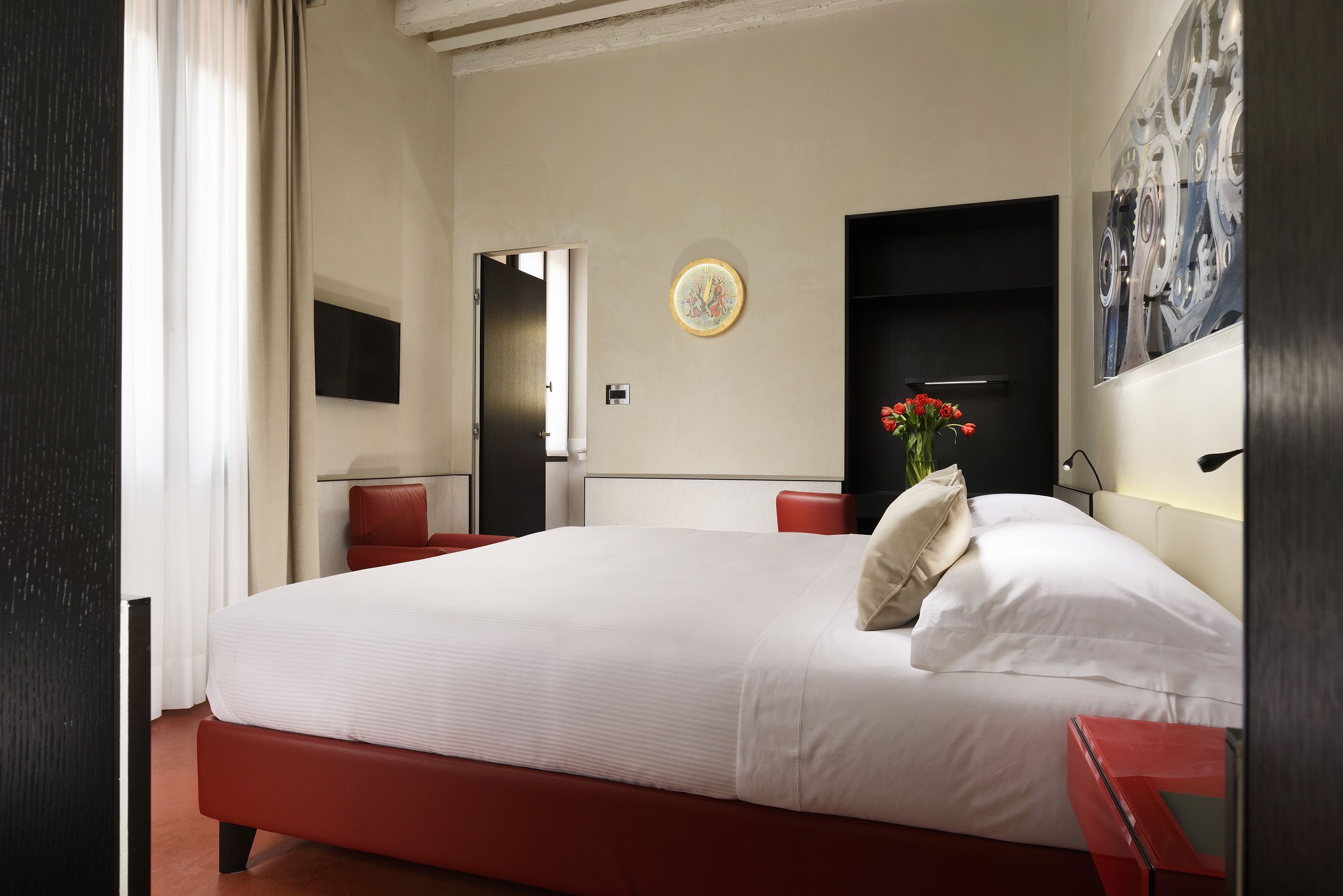 A hotel room with a large bed, a red chair, a small table, a wall-mounted TV, and a painting.