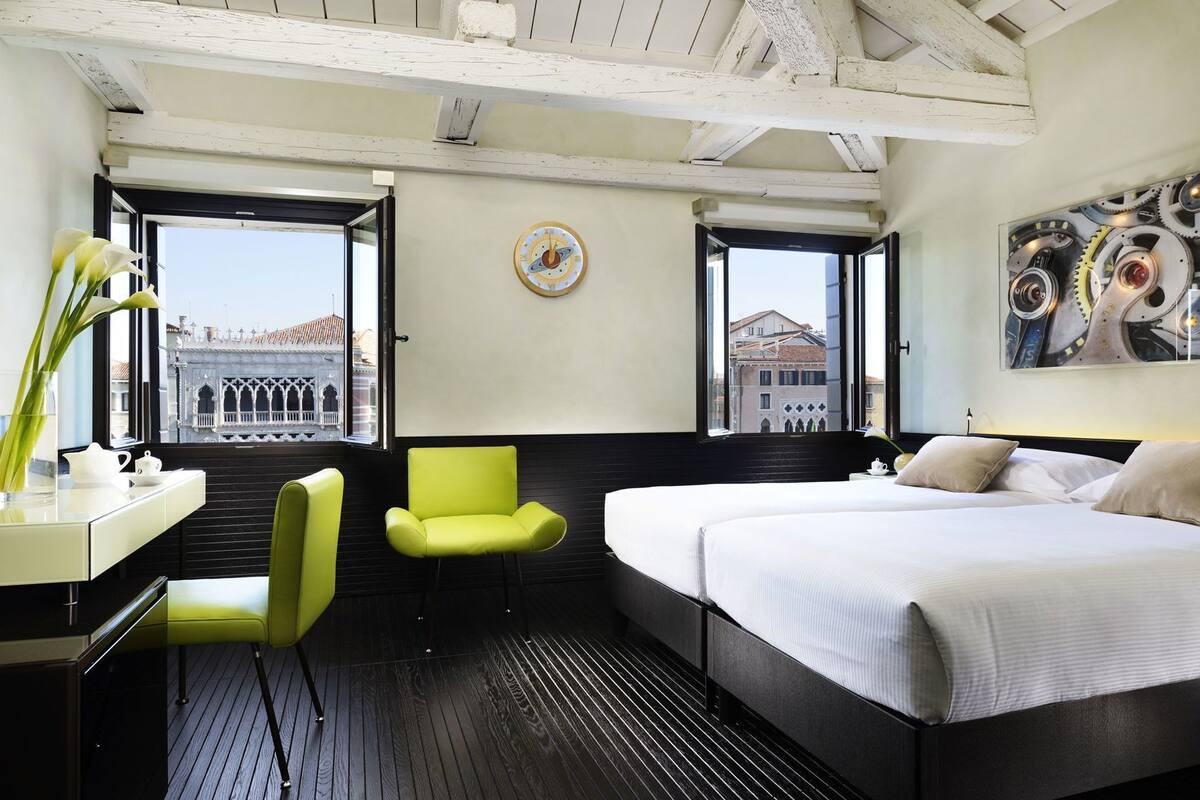 A hotel room with a bed, a desk, a chair, and a large window with a view of buildings.