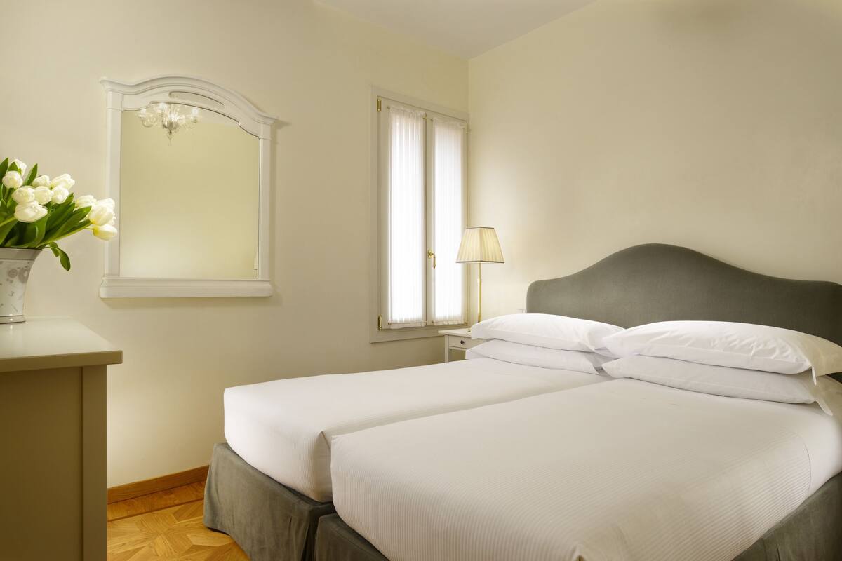 A hotel room with two beds, a bedside table, a lamp, a mirror, and a window with curtains.