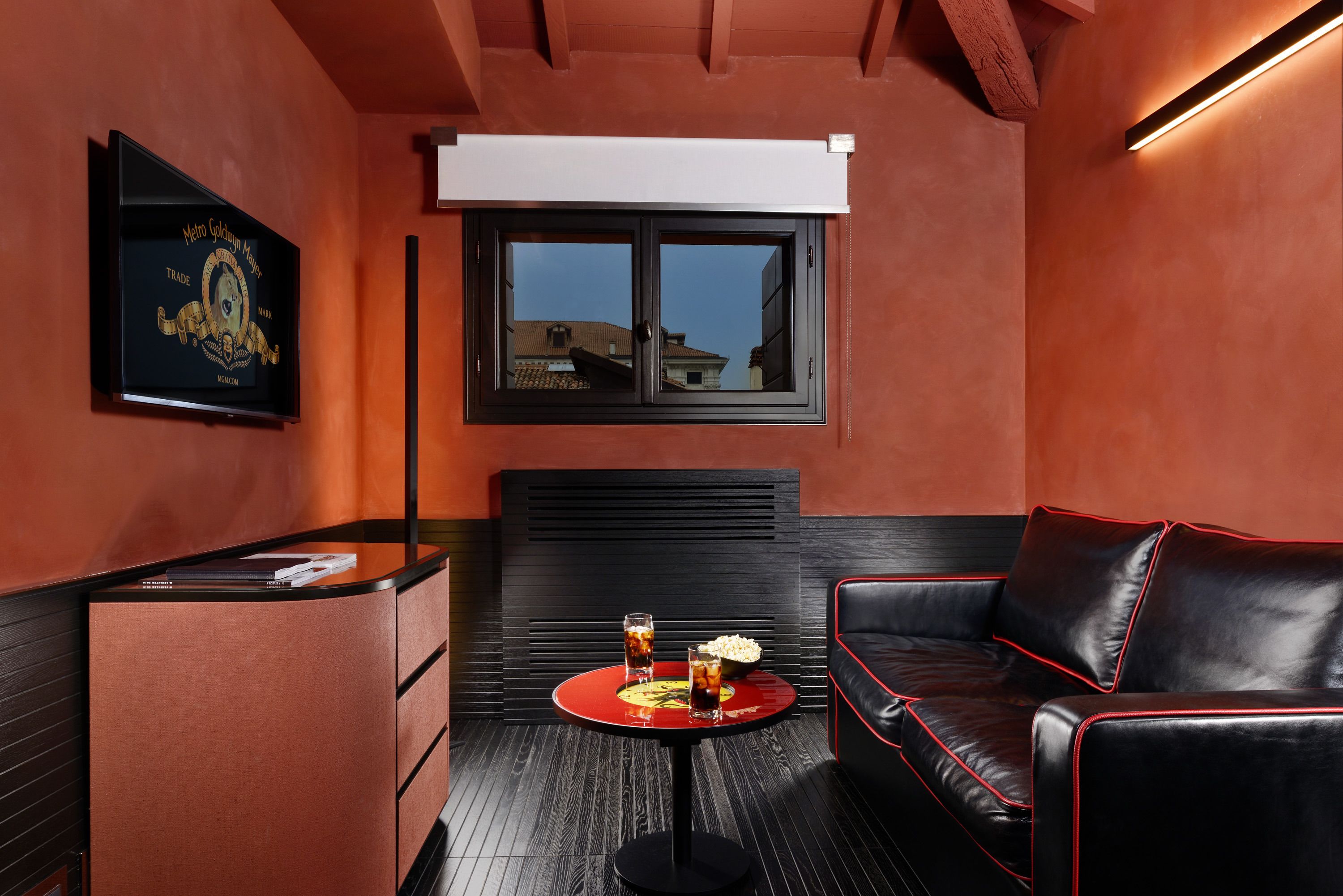 Una habitación con sofá de cuero negro, una mesita redonda con bebidas, un minibar, una ventana con persianas y un cuadro en la pared.