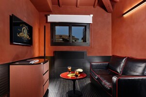 A room with a black leather sofa, a small round table with drinks, a minibar, a window with blinds, and a framed picture on the wall.