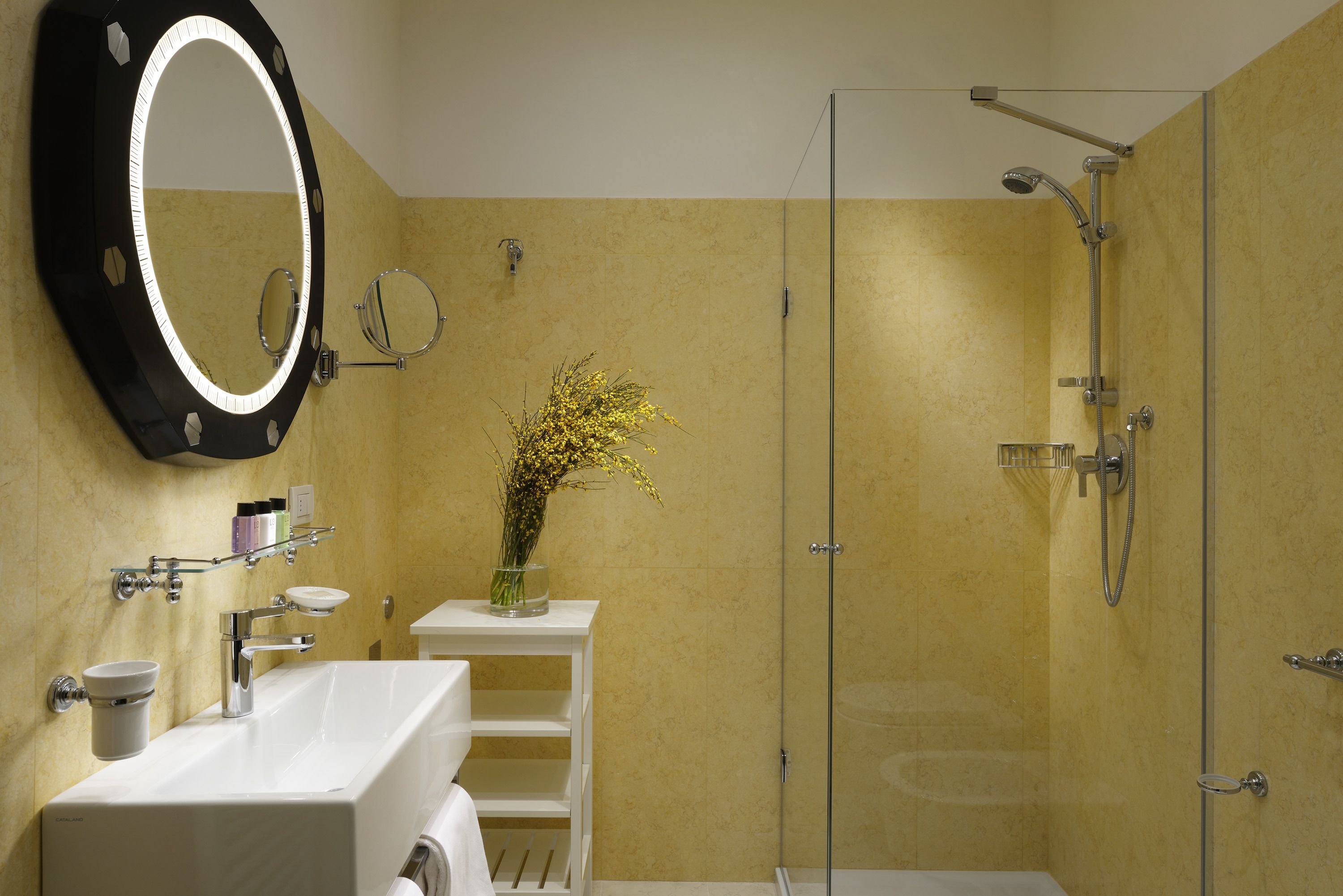 A bathroom with a large mirror, a white sink, a glass shower enclosure, and a vase with yellow flowers on a small shelf.